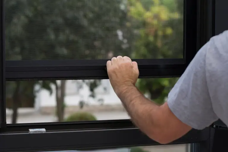 Mano bajando la barra de una mosquitera enrollable en una ventana de perfil negro con jardín detrás